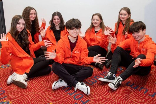Group of smiling teenagers wearing matching bright orange hoodies, seated on a red patterned carpet, posing together with hands raised in a fun and welcoming gesture.