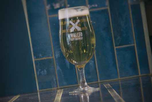 A pint glass of beer with a foamy head, branded with the "Walled City Brewery" logo, placed on a reflective bar counter with a blue tiled wall in the background.