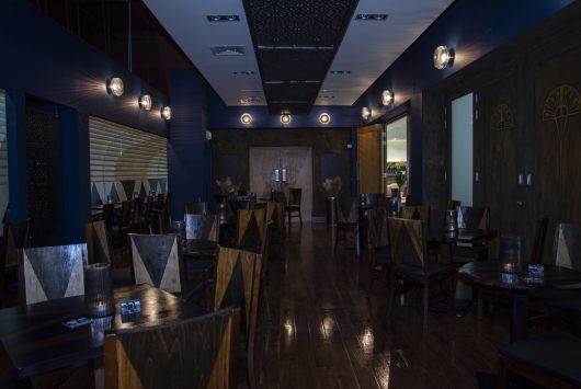 A dimly lit modern Speakeasy bar with dark wooden floors, navy walls, and neatly arranged triangular-back chairs around small tables, creating an intimate and stylish atmosphere.