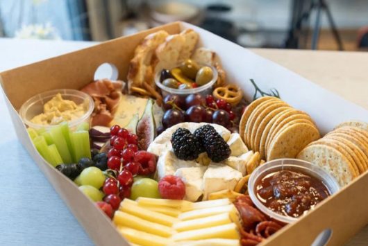 A gourmet charcuterie box filled with an assortment of cheeses, crackers, cured meats, fresh grapes, sliced fruit, dips, and berries, arranged neatly in a kraft tray on a countertop.