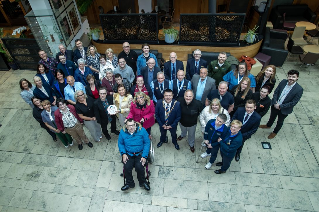 Overhead group photo inside the Millennium Forum foyer showing staff members, accessibility advocates, community organisations and local representatives gathered together to celebrate the venue receiving Accessibility Champion Status. A wheelchair user is positioned at the front of the group while others stand across the large tiled marble floor smiling towards the camera. The bright modern foyer features glass display cabinets, wooden panelling, black decorative screens, indoor plants and seating areas in the background.