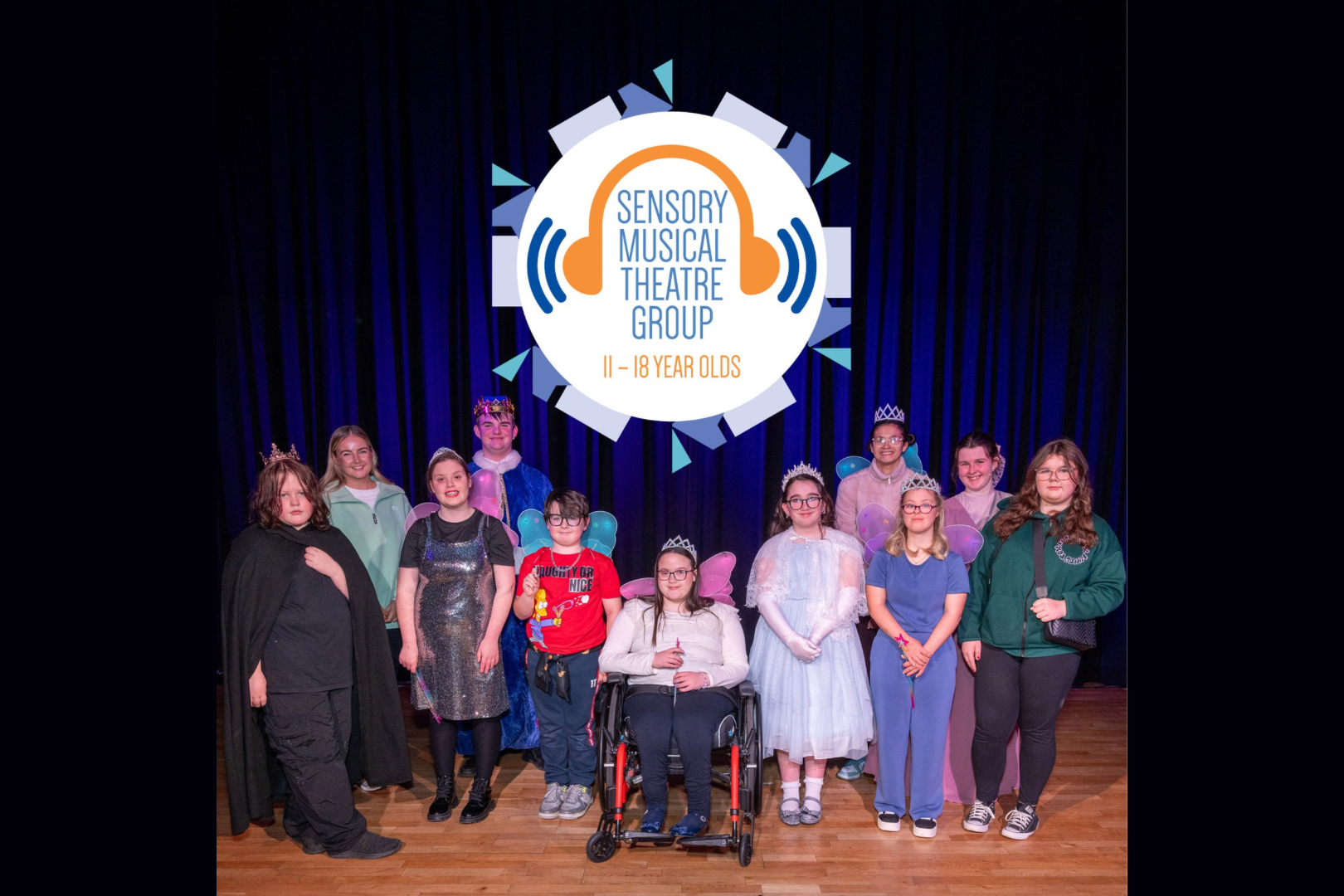 A group of teenagers stand on a stage in front of a dark blue curtain, posing for a photo beneath a large circular logo that reads “Sensory Musical Theatre Group – 11–18 Year Olds.” The young people wear a mix of casual clothes and costume accessories such as crowns, fairy wings, and sparkly outfits, with one person seated in a wheelchair at the center. The stage floor is wooden, and the group faces the camera smiling.