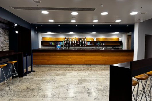 A modern East Wall bar area with a long wooden counter, a selection of beverages on display, high stools along the sides, and bright ceiling lights illuminating the marble-tiled floor.
