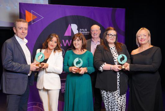 Six people stand in front of a purple Arts & Business NI Awards backdrop, with four holding glass awards and smiling for the camera.