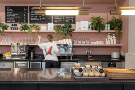 Een vrouw maakt een drankje achter de counter van een café, met een roze muur achter de apparaten en planten en servies uitgestald op witte planken.