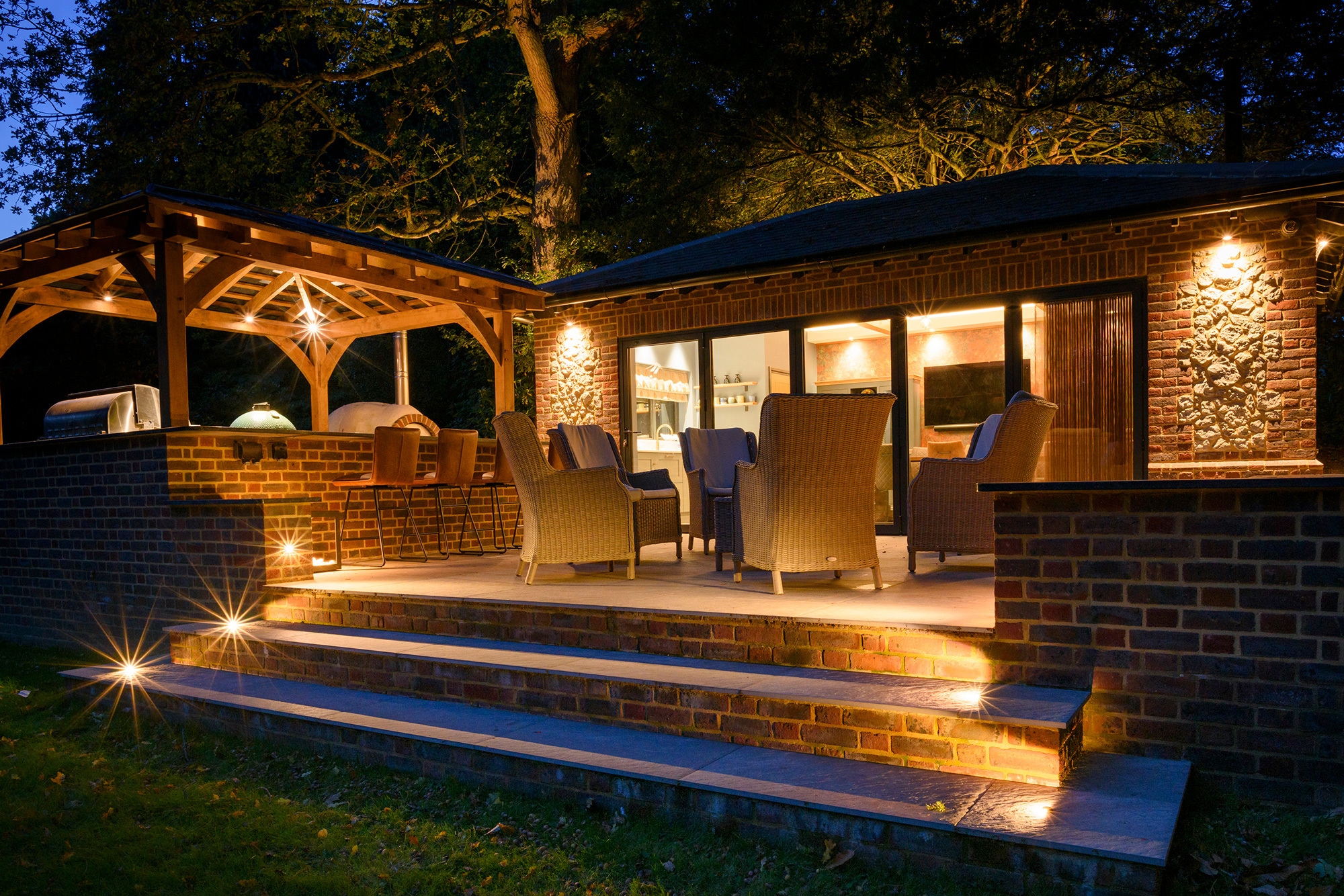 Brick terrace and oak pavilion at dusk with warm step lights, wall lights and lit outdoor kitchen