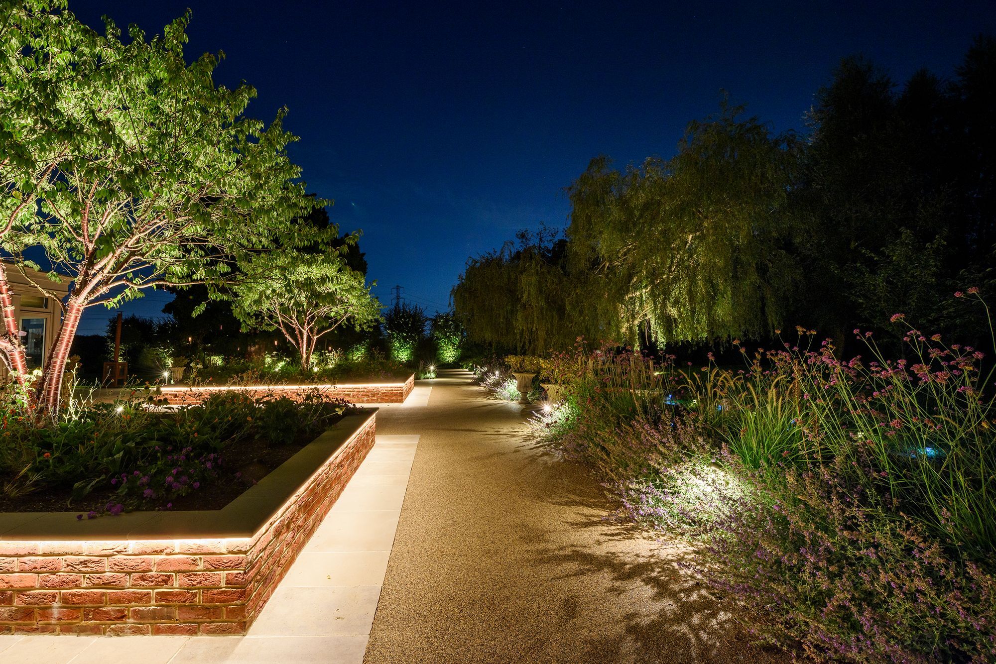 Raised brick planters with warm LED strip lighting beside a resin bound path and uplit trees at dusk in Denham