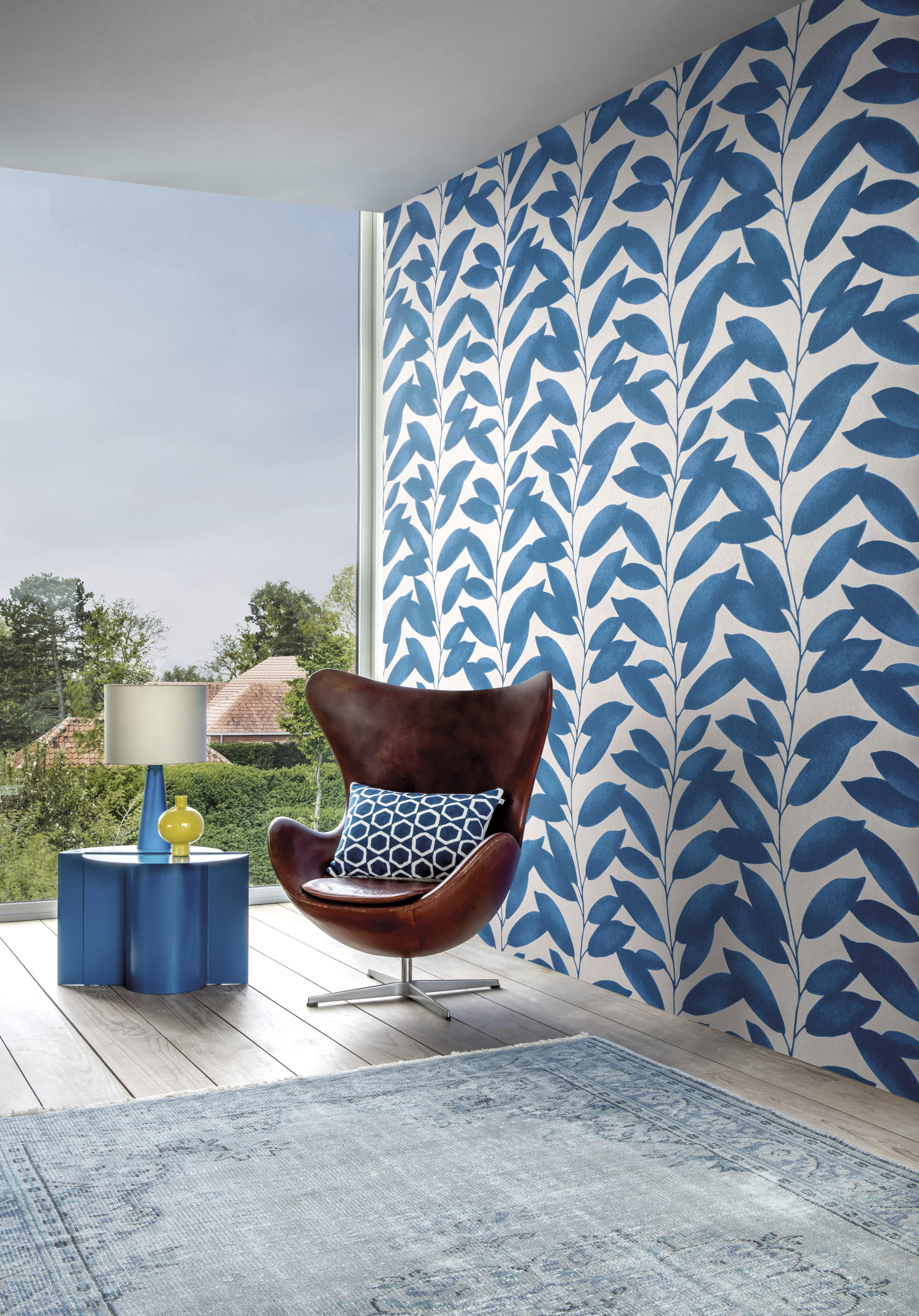 This image shows the Blooming Marvellous design from Omexco's wallcoverings collection Joy. The playful design with a pattern of blue leafs is the perfect backgrop for the iconic Egg chair designed by Arne Jacobsen. The large window adds brightness to the contemporary space.