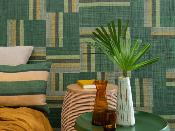 Authentic craftmanship embodies the bold Atelier collection designed by the creative minds of Omexco. This image shows an impressive green patchwork of handwoven raffia. The perfect statement wall in this tropical bedroom with ochre colour accents, a green metal bedside table, exotic plant leaf and cosy bed pillows.