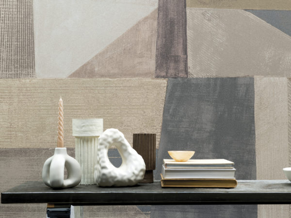 Moonstone by Omexco is a lavish collection of textured wallcoverings softly iridescent like the Moonstone. In this picture we see a feature wall with an XXL abstract art printed on fine mica. The geometric design with diagonal lines is colorful, yet not overwhelming with its soft shades of beige, white, grey, soft pink and unsaturated blue. In front of the wall, we see a low wooden coffee table with small vases and other ceramic decorations as well as a pile of books.