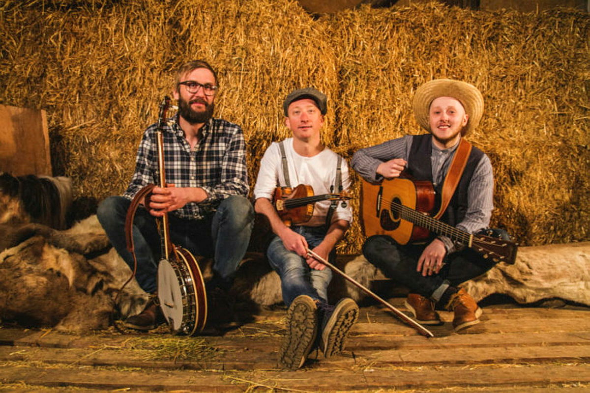 Tre musikere med banjo, fiolin og gitar sitter på høyballer i en rustikk låve.