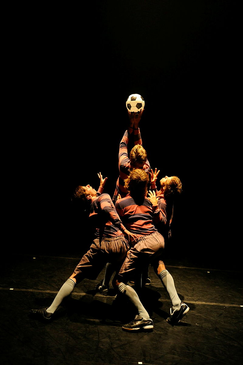 Utøvere gjenskaper et dramatisk fotballøyeblikk på scenen, og strekker seg etter en opplyst ball mot mørk bakgrunn i teatralsk positur.