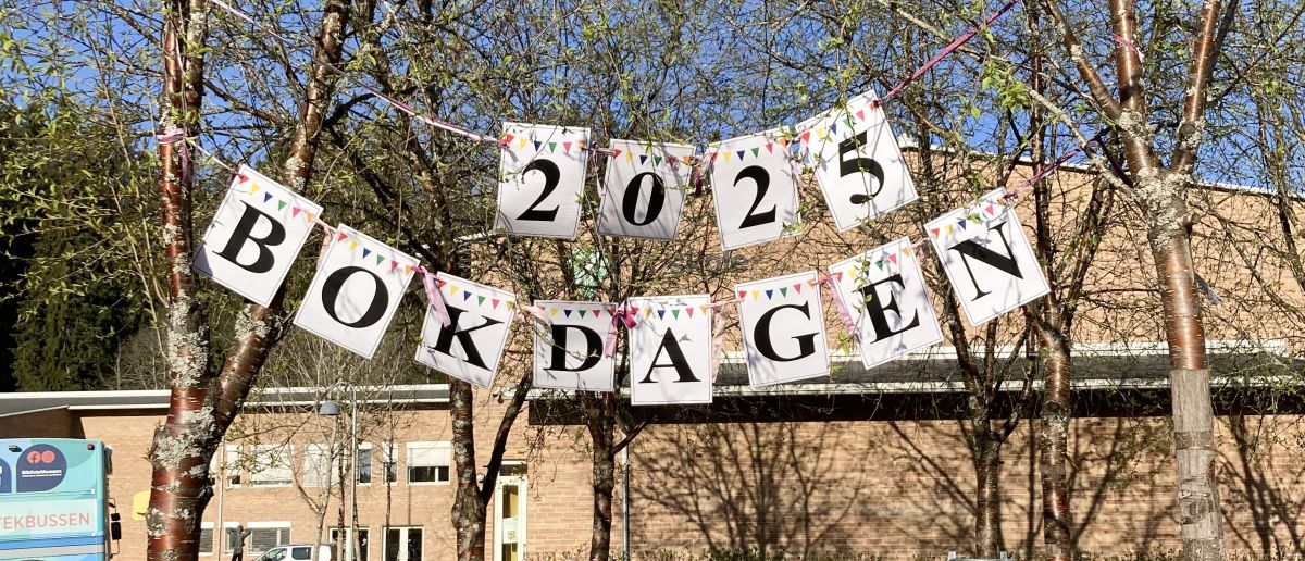 Det er sol og vår, og et banner henger mellom to trær. På banneret står det skrevet 2025 Bokdagen.
