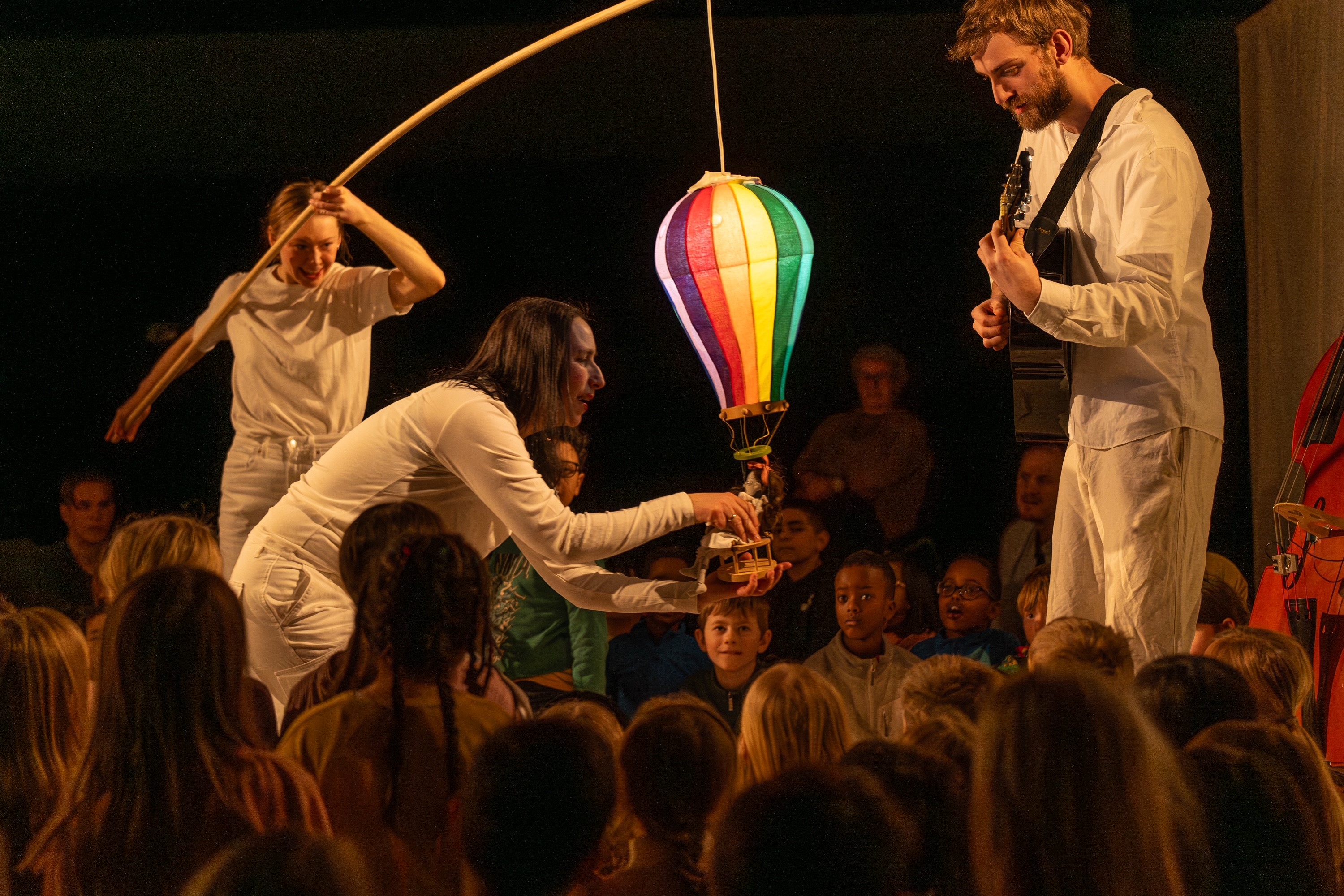 Utøvere engasjerer barnepublikummet med fargerik luftballongrekvisita. Mannen spiller gitar, kvinnen samhandler med ballongen, barna ser nøye på.