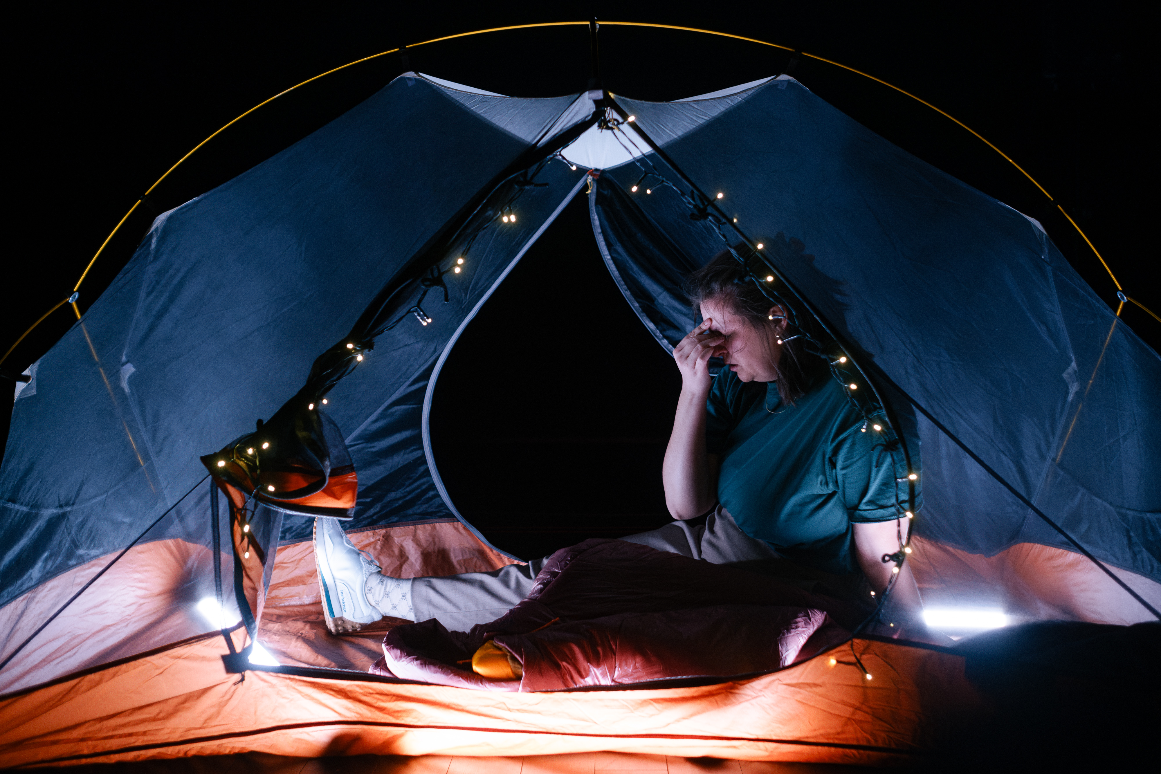 En ung jente sitter inni et telt med gjennomsiktig teltduk med rød bunn, og en lyslenke som lyser rundt åpningen på teltet. Hun ser sliten ut, gnir seg i øynene og er svett i håret.