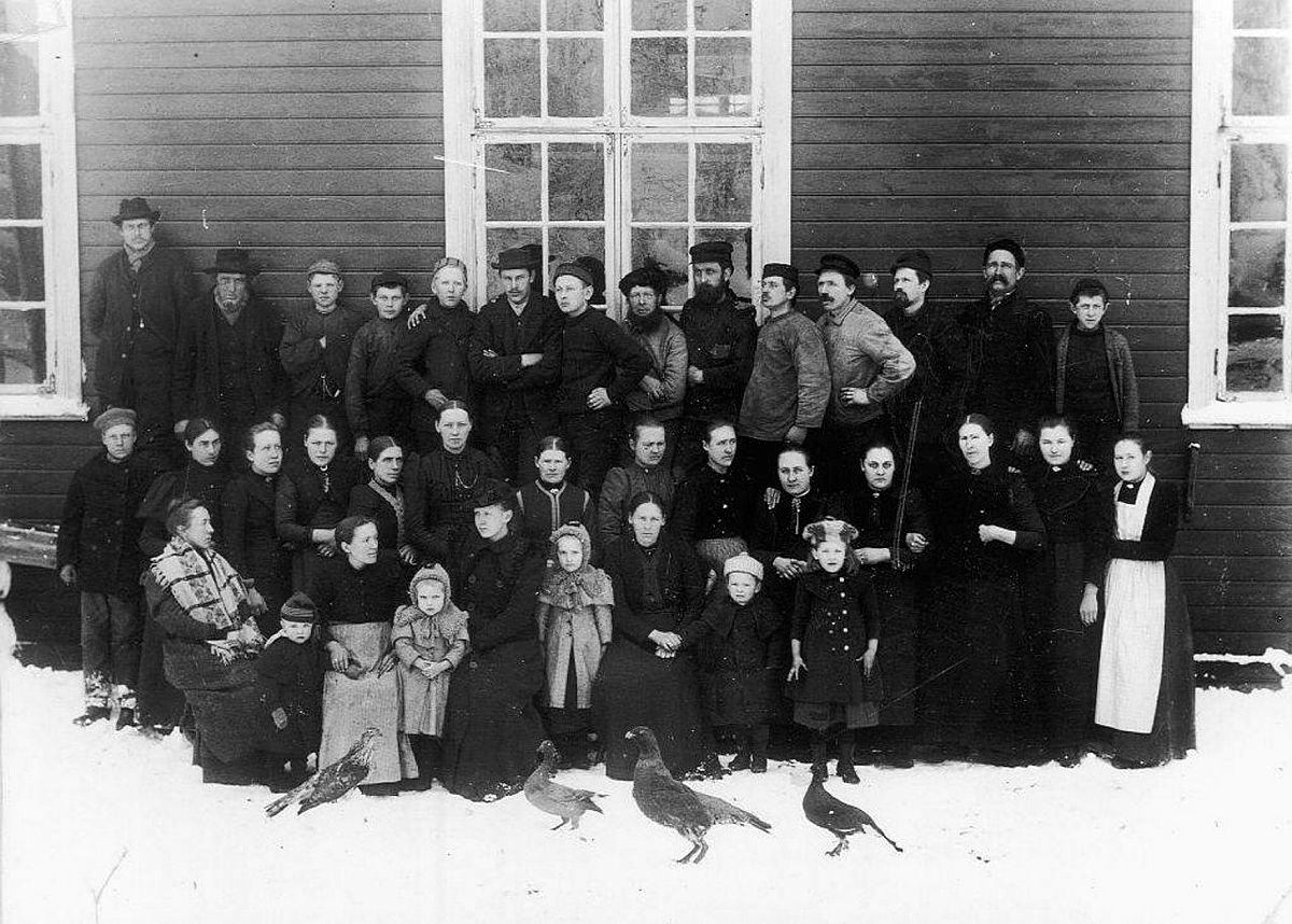 Ansatte ved fabrikken i 1898, stilt opp foran en eldre fabrikkbygning i tre med store, sammensatte vinduer. Kvinner og barn i de to fremste rekkene, og menn i den bakre. Alvorlige ansiktsuttrykk. Malplasserte fugler i fremkant.