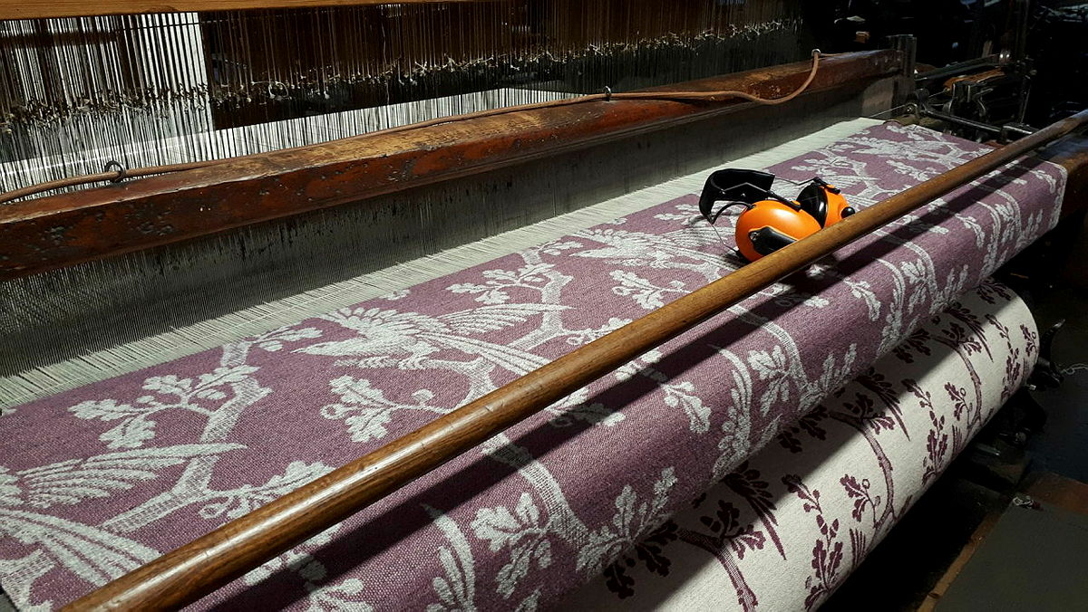 Utsnitt som viser deler av en gammel vevstol. Stoffet på bildet blir vevd etter et hundre år gammelt mønster, og motivet viser en fugl som sitter på en gren i et tre med eikeløv. Fargene er duse.