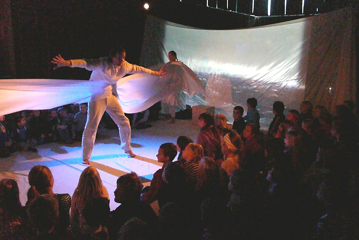 Utøver i hvitt antrekk på scenen med utstrakte armer, underholdende sittende publikum. Stort hvitt bakteppe bak. Dramatisk belysning.