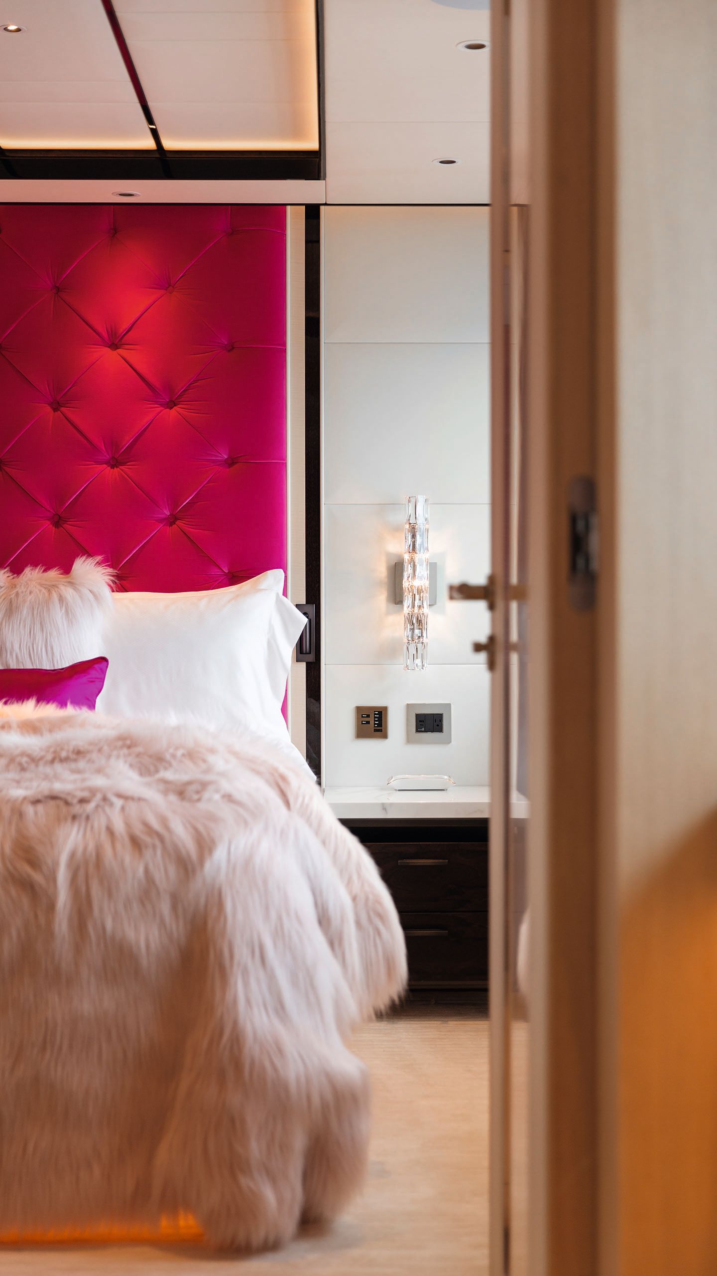 Close-up of a bed which has a bright pink headboard and pink cushion, furry throw and cushion