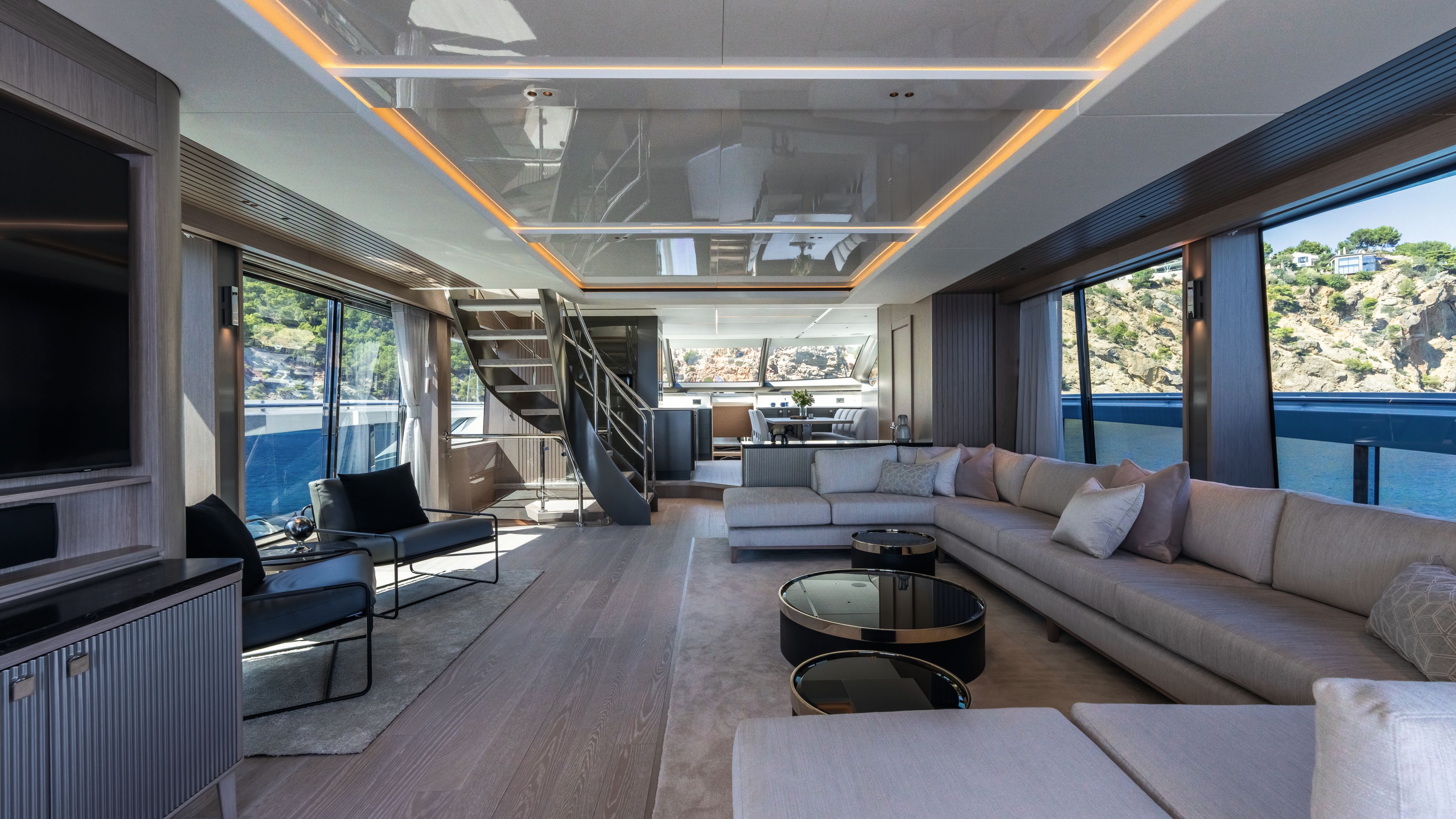Main deck showing large U-shape leather sofa, soft chairs, spiral staircase and round black coffee tables with TV to one side on the wall