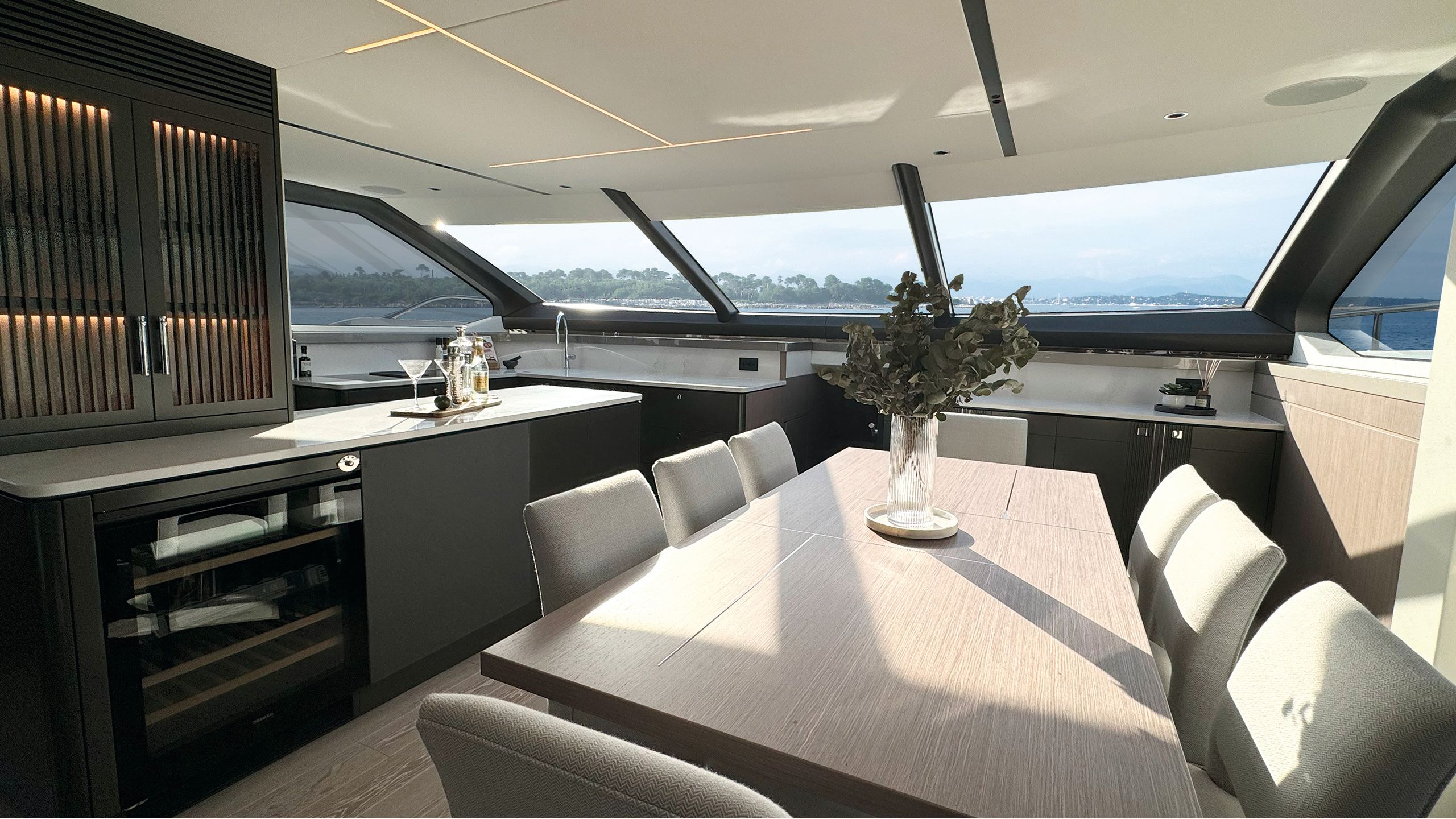 Dining table with seating for 8, under a slanting window. The counters are white marble and the cabinets are dark. A bunch of eucalyptus is in a ribbed clear vase on the table and there are martini glasses and cocktail shakers on the counter.