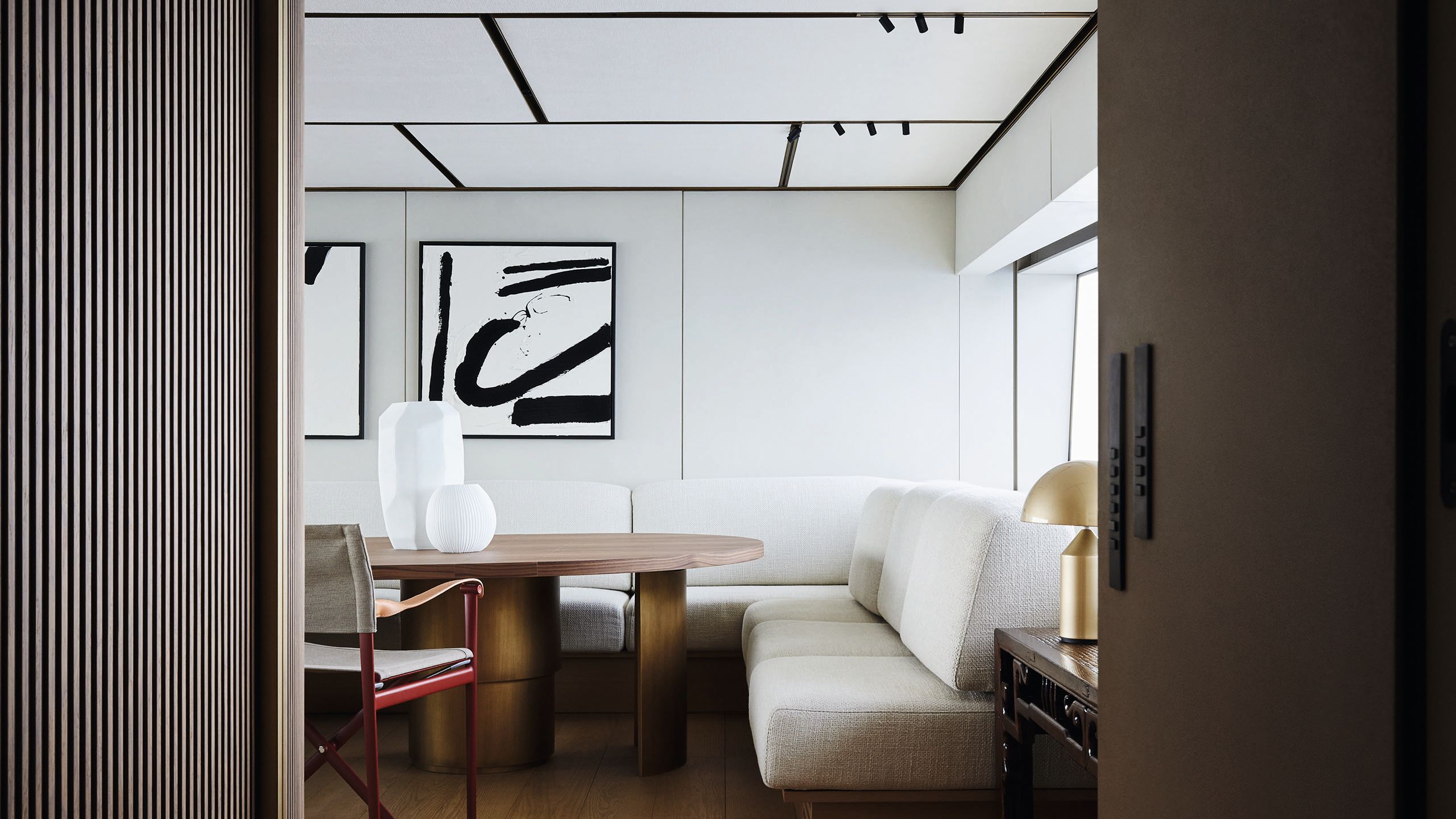 Looking into the media lounge from a different angle. There is abstract white and black art on the walls, a table with geometric white bases and white L-shaped sofas. The wall features ribbing in dark wood