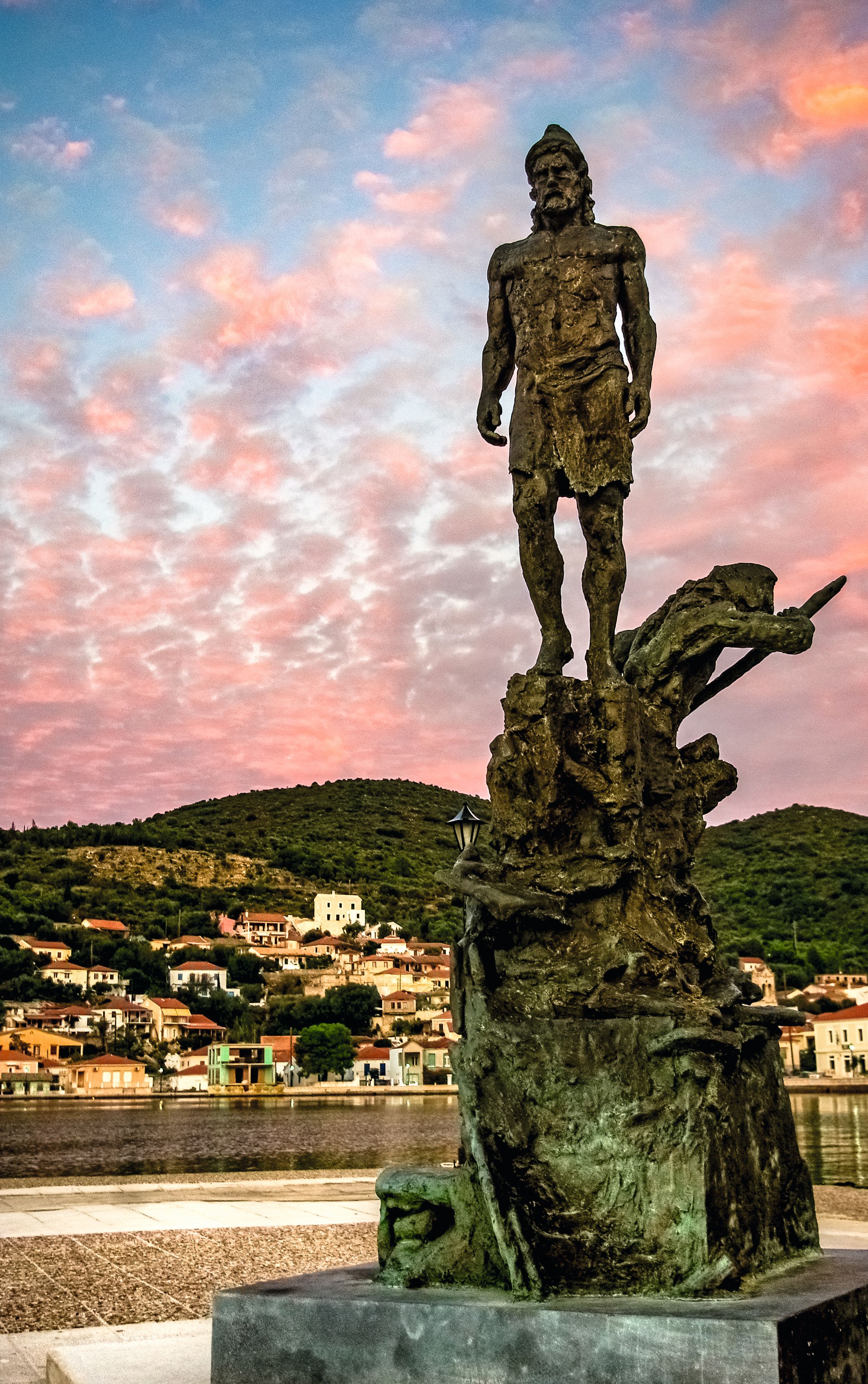 A statue of Odysseus - it is on a large stone plinth. Ithaca is behind and the sky is blue with pink clouds