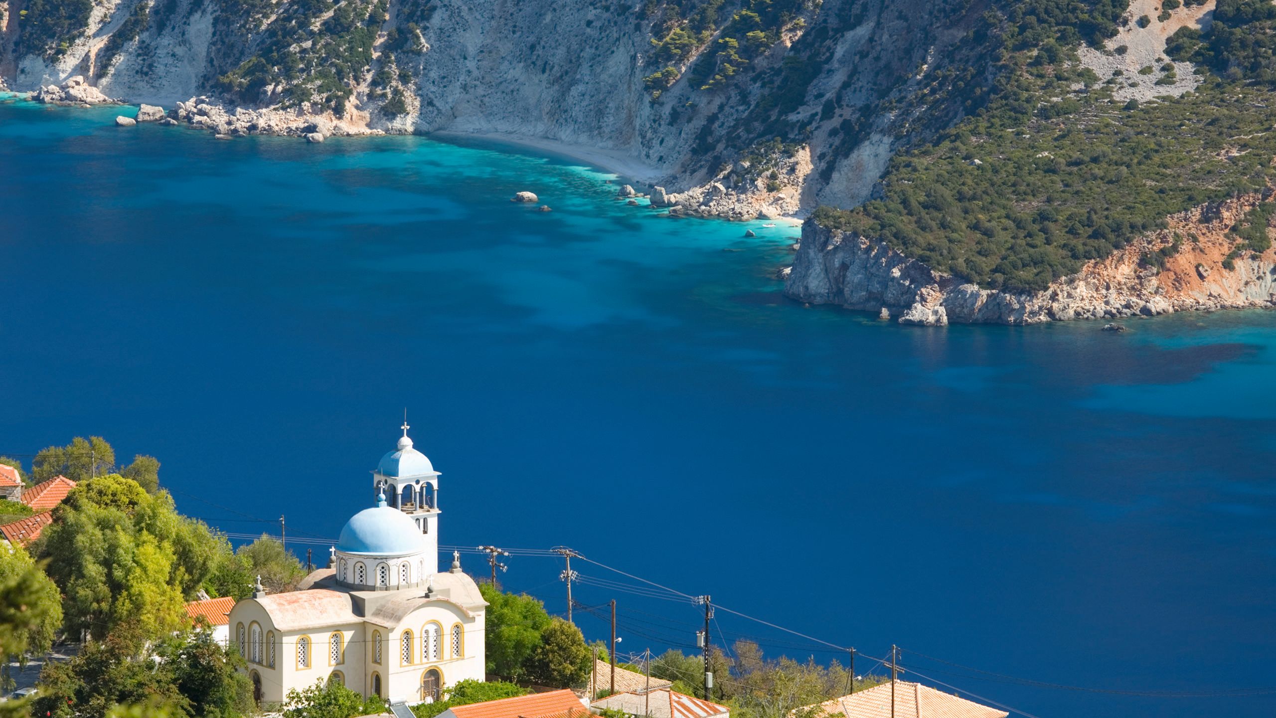 Corfu from above. There is a blue-domed white church in the foreground perched on a clifftop, looking out on to deep blue waters