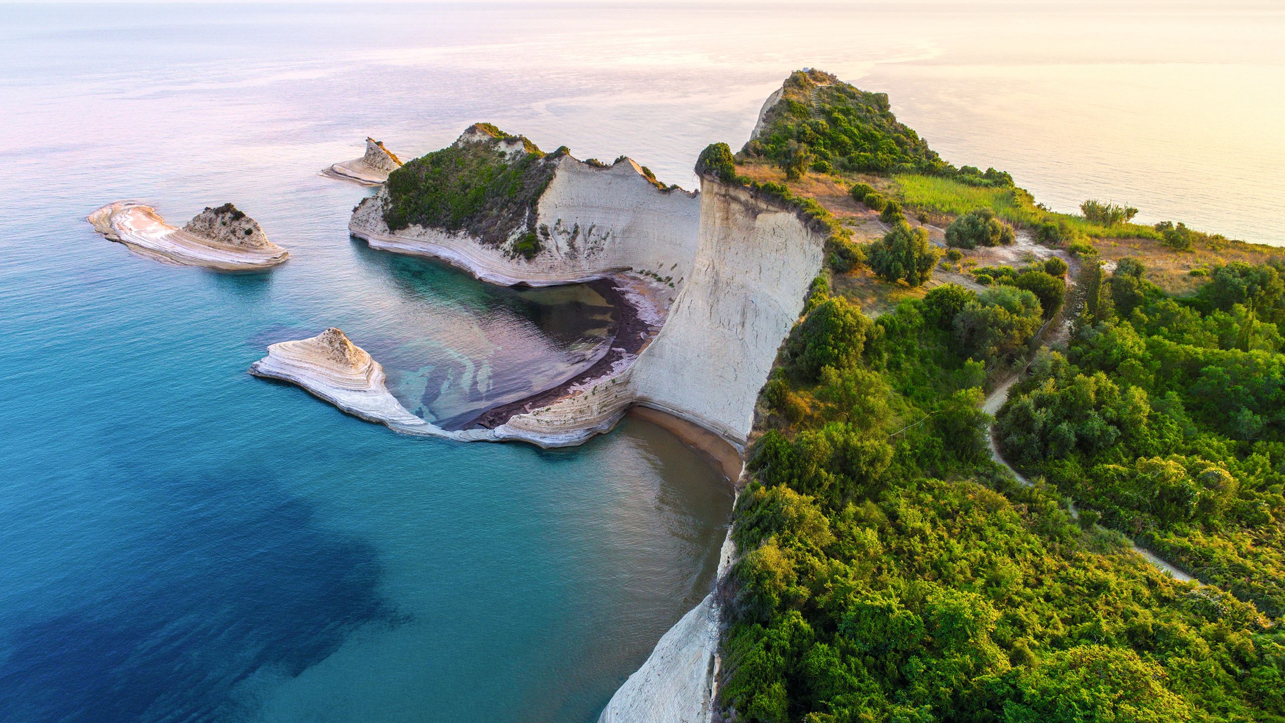 Corfu's coastline from above, focussing on white-sided cliffs topped with greenery. The bay in front is turquoise and dark blue