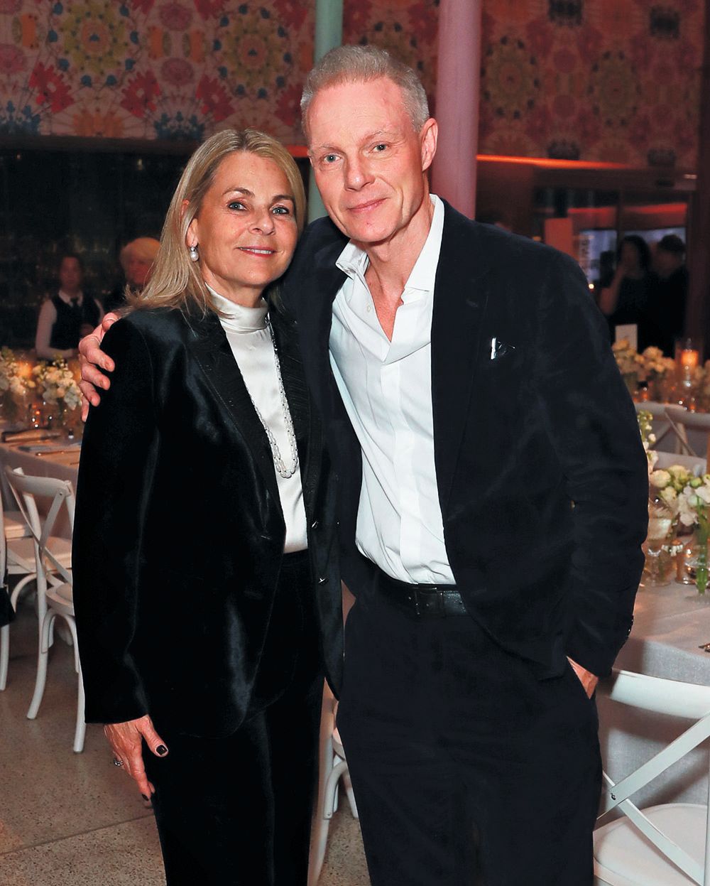 Reuben in a two-piece black suit and white top. A man in a black suit has his arm around her