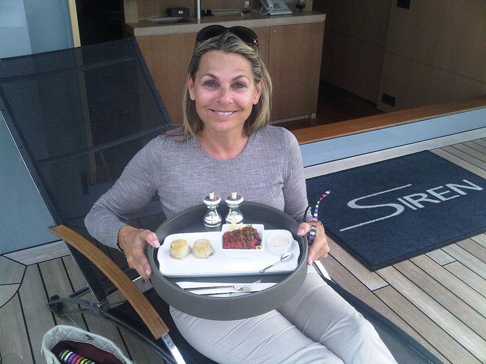 Debra sitting in a chair holding a grey tray that has a white plate of food on it. She has a dark blonde bob, sunglasses on her head and is smiling. She is wearing a grey long sleeve top and light trousers