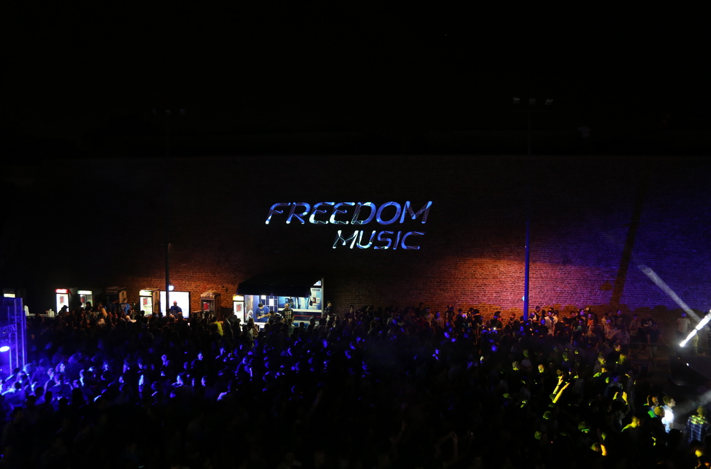 A crowd of concert-goers illuminated by neon lights, with the words "FREEDOM MUSIC" projected in large, floating letters on the brick wall above the event entrance.