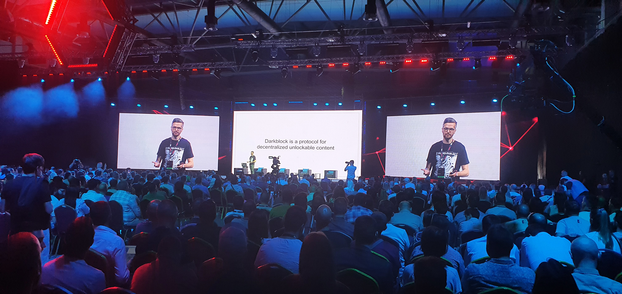 A speaker at the TMRW Conference presents on the topic of decentralized unlockable content, with a large attentive audience and a backdrop of the conference's branding and futuristic red and blue lighting.
