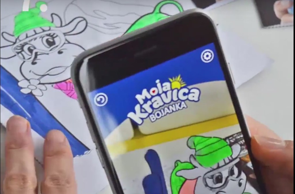 Close-up of a smartphone using the 'Moja Kravica' app to overlay a colorful, augmented reality image onto a child's coloring book page, creating an interactive experience.