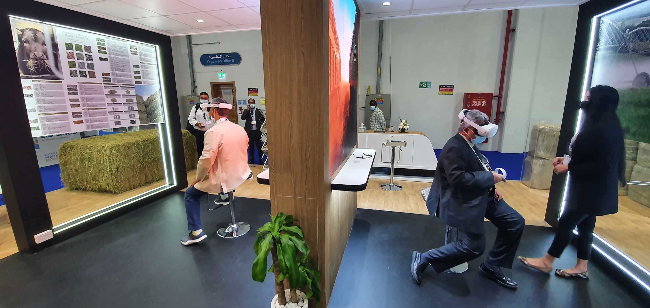 Two people are engaged in a virtual reality demonstration in a busy exhibition space.