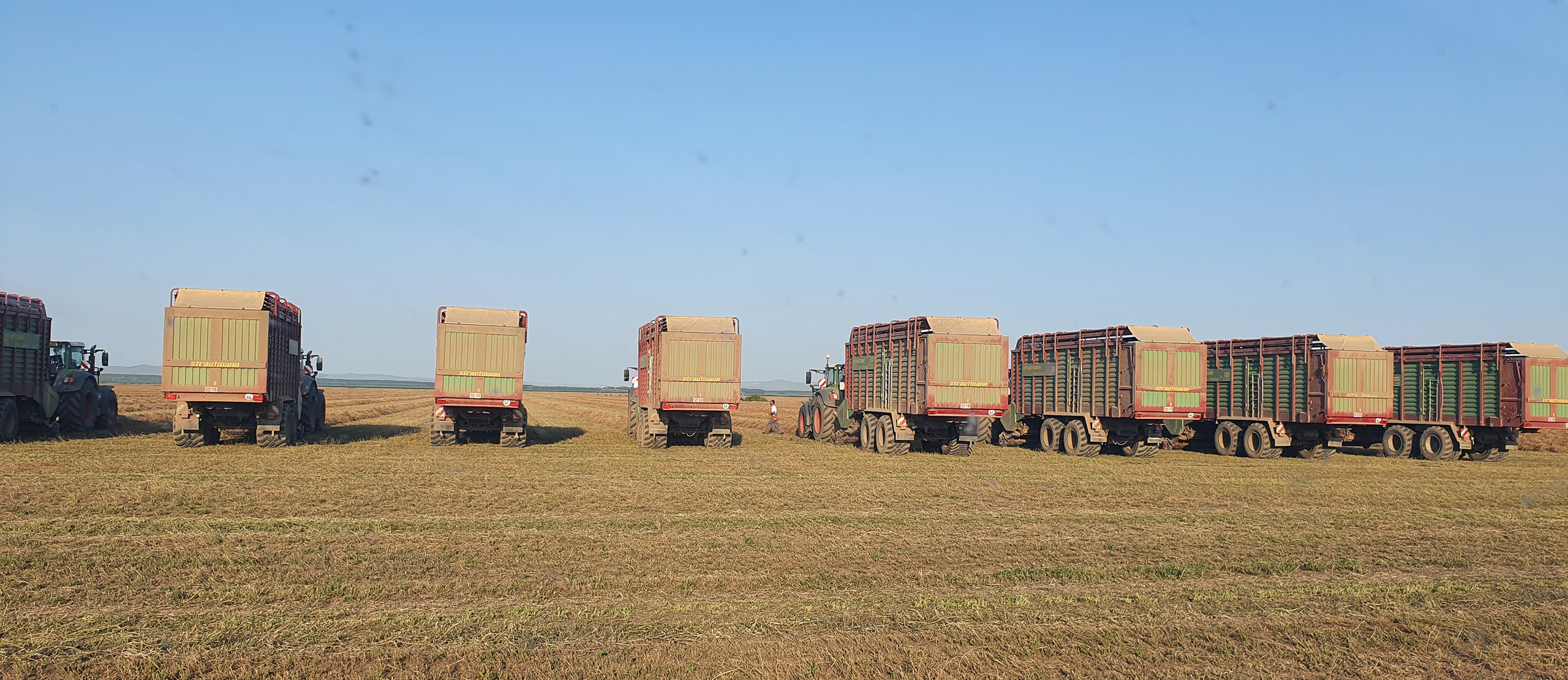 An expansive view of agricultural land with multiple large balers lined up in the background, ready for fieldwork. The landscape is flat and vast under a clear sky, with a slight haze near the horizon.