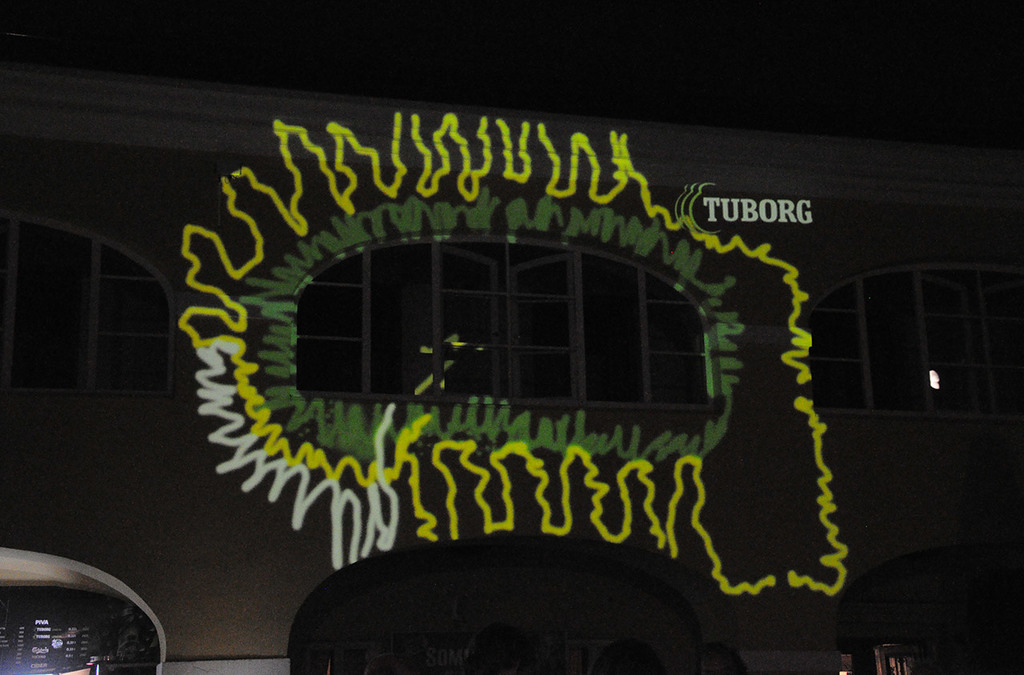  A building facade is illuminated by a green, laser-painted pattern resembling a splat, with the word "Tuborg" projected above. 