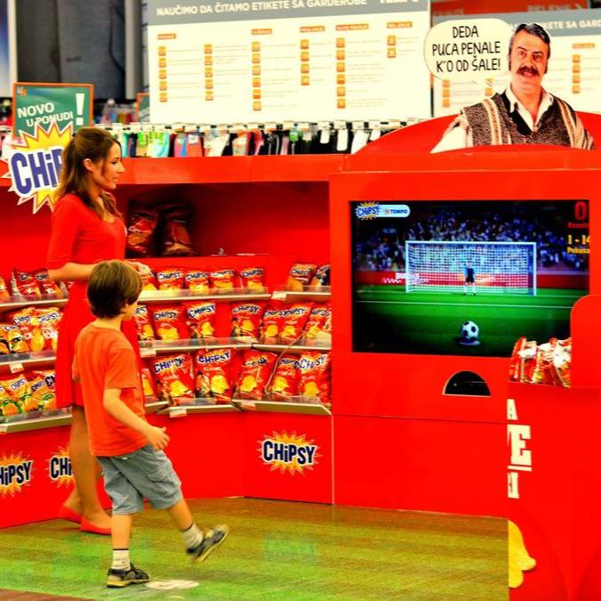 Shoppers in a grocery store pass by a promotional setup for Chipsy, featuring a soccer penalty kick interactive game, blending retail with entertainment.