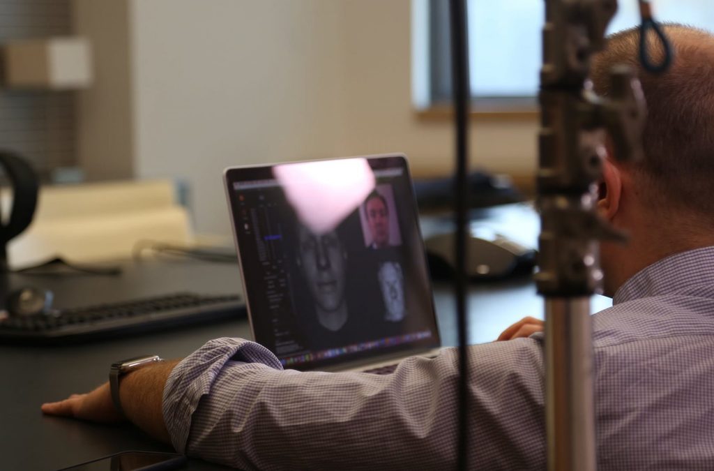 Behind-the-scenes view of a workspace with a computer screen showing the 3D modeling process of the GM's digital avatar.