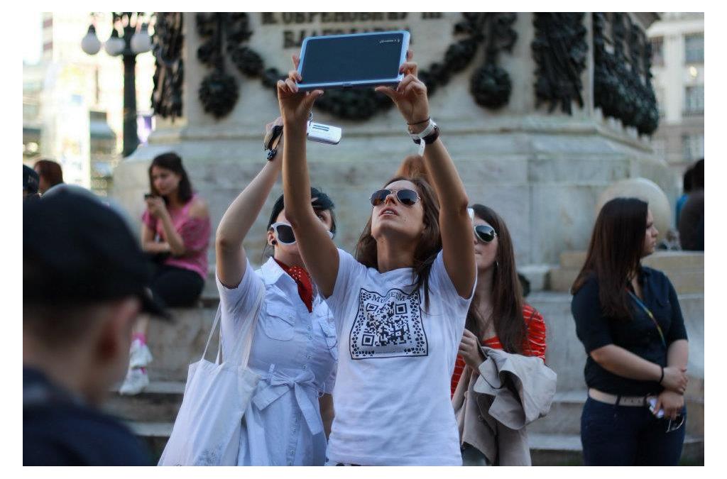 A person uses a tablet to scan a special ARQR code from the MUZZEUM project displayed on a building, revealing a virtual artifact on their screen.