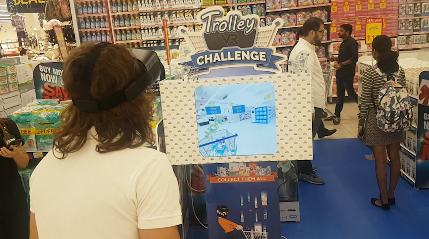 A participant is engrossed in the 'VR Trolley Challenge', wearing a virtual reality headset and standing in front of a promotional display in a supermarket. The screen above shows a 3D shopping environment where the game takes place, set against a backdrop of busy supermarket aisles.