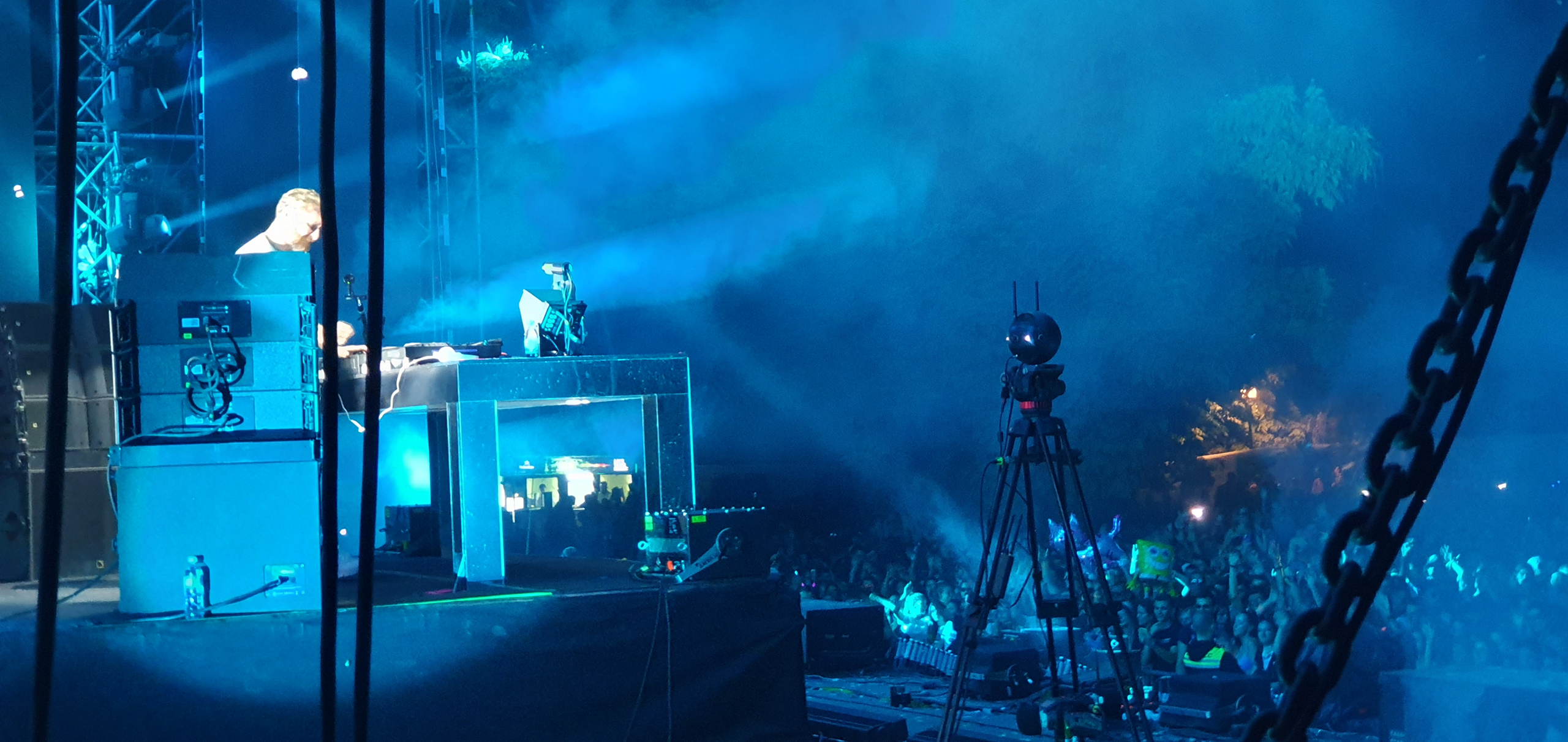 A side-stage view of a music concert showing audio equipment and a DJ performing, with blue stage lights casting a hazy glow over the scene. In the foreground, various cables, equipment, and the silhouette of a large chain suggest backstage access. A crowd is partially visible in the background, immersed in the event.