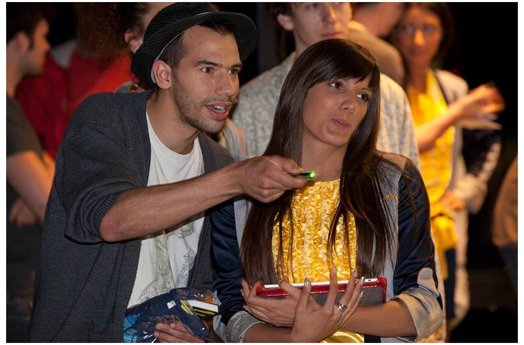 A man holding a laser pointer participates in a laser painting session, as a woman beside him watches intently, holding an iPad. They are interacting with the projected surface, exploring the novel digital art technique.