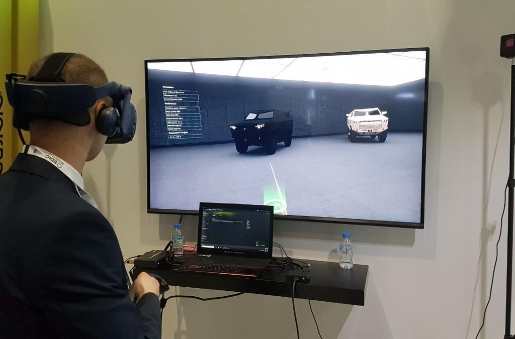 A visitor at IDEX 2019 engaging with a virtual reality presentation of the Despot 4x4 vehicle using an HTC Vive Pro headset, with the digital model visible on the monitor and a brochure visible in the foreground.