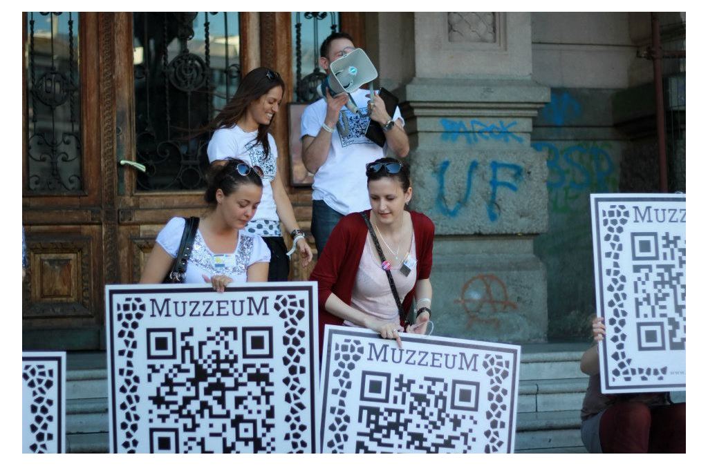 People participate in the MUZZEUM project outdoors by holding and scanning ARQR codes, accessing 3D representations of cultural heritage items on their mobile devices.