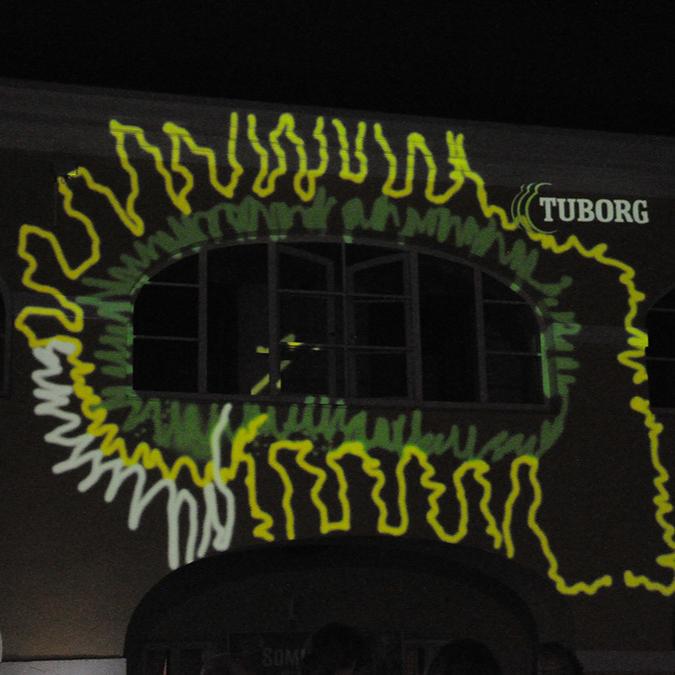  A building facade is illuminated by a green, laser-painted pattern resembling a splat, with the word "Tuborg" projected above. 