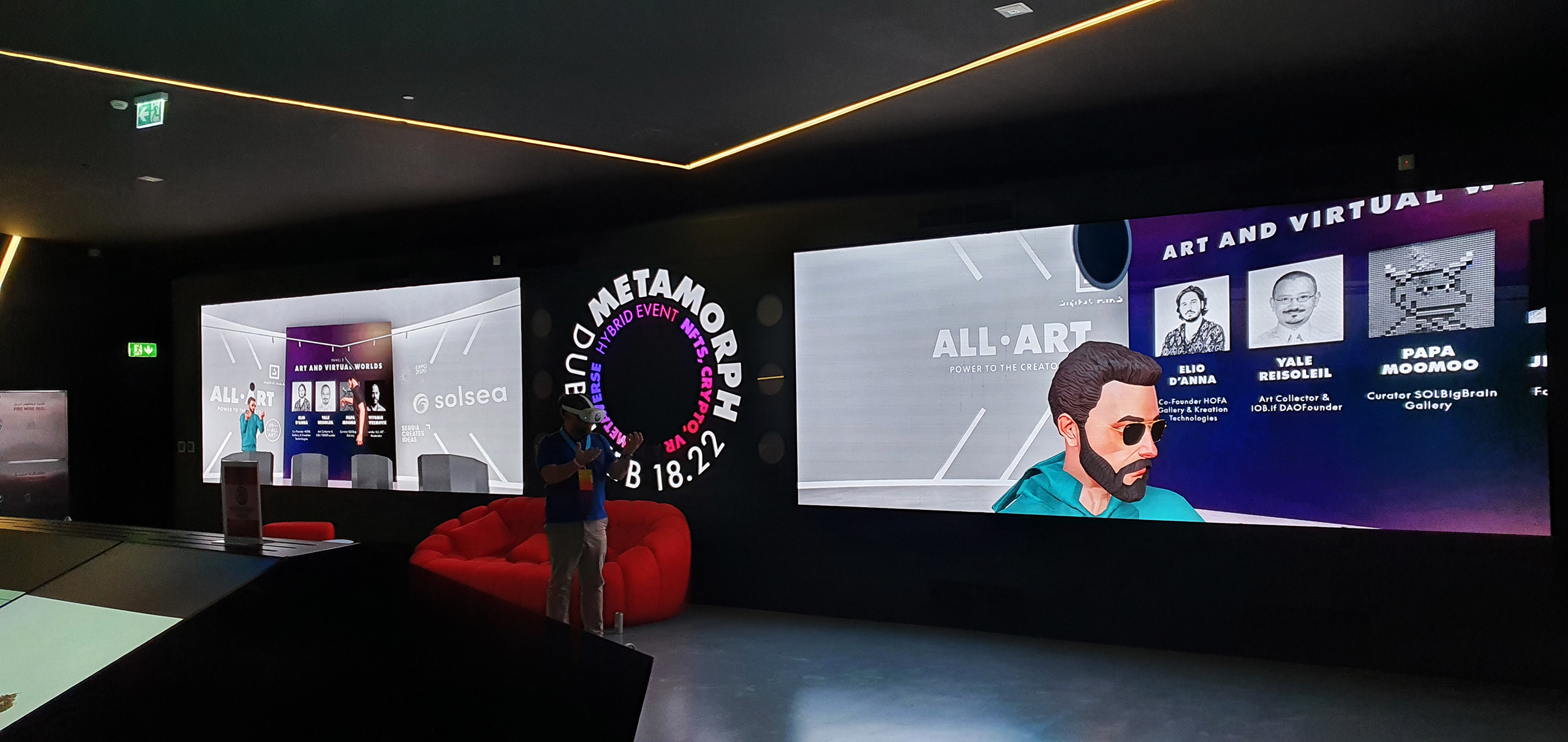 The interior of the Metamorph event venue displaying an immersive space with a modern, dimly lit lounge area. Monitors display the event’s branding and participant avatars, emphasizing the hybrid nature of the conference that combines live interactions with virtual reality experiences.