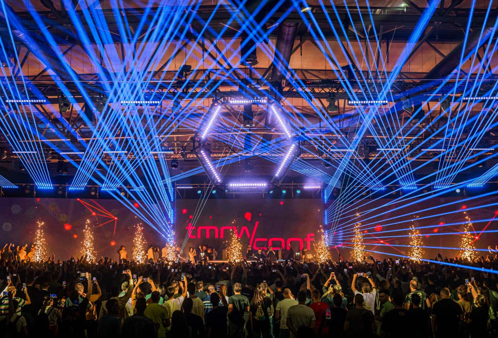 A vibrant concert atmosphere at the TMRW Conference in Belgrade, showcasing an elaborate light show with dynamic blue lasers crisscrossing above the crowd.