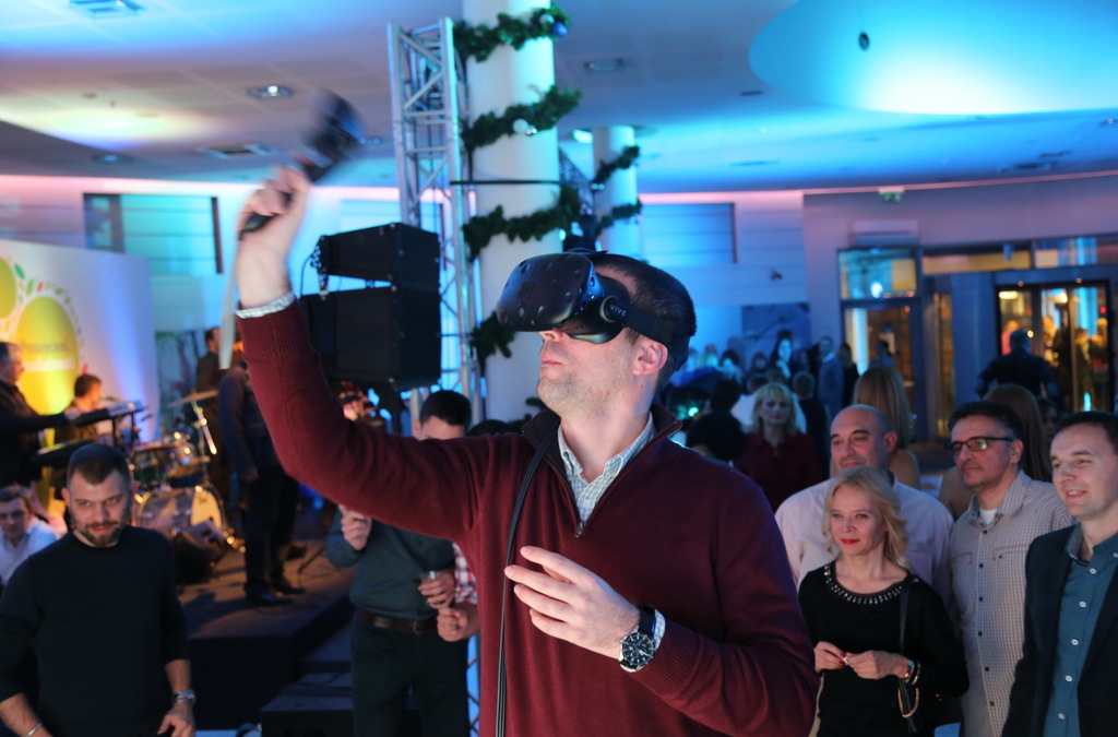 A man in a red sweater and VR headset enthusiastically raises his controller, simulating a basketball shot in a lively event atmosphere with spectators in the background.