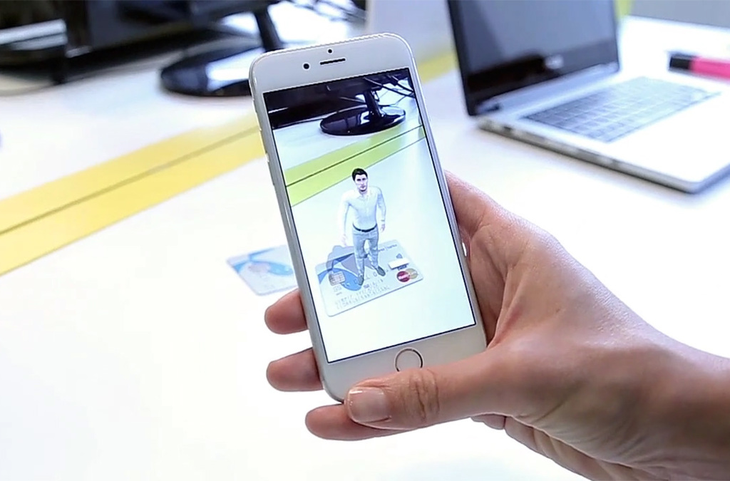 A person's hand holding a smartphone displaying an augmented reality (AR) image of a digital banker standing on a Telenor Bank card. The digital banker is gesturing welcomingly within the screen of the smartphone, which is being viewed over a desk with a laptop in the background.